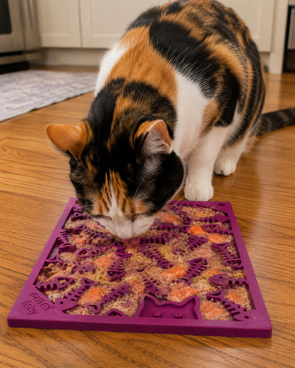 A calico cat licks food from a small purple SodaPup Fishy Design EMat enrichment lick mat with varied patterns on a wooden kitchen floor, keeping the cat engaged during mealtime.