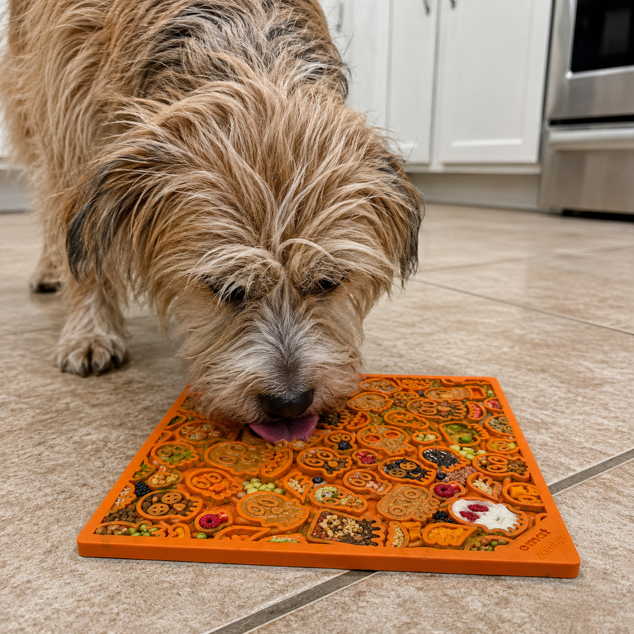 A scruffy dog enjoys treats from the SodaPup Zombie Design Emat Enrichment Lick Mat on a tiled kitchen floor, with white cabinets and a stainless steel oven in the background.