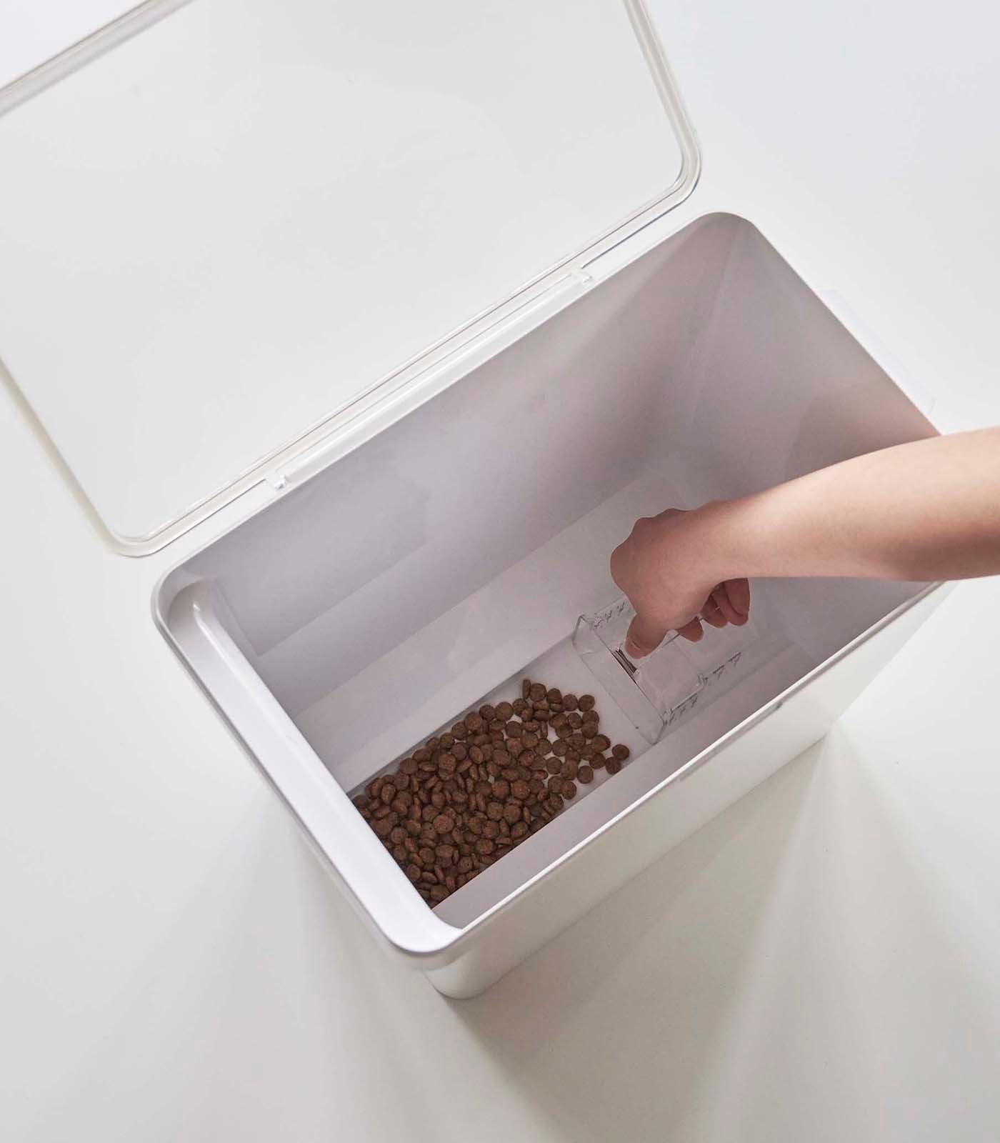 A person's hand is pouring dry pet food from a clear measuring cup into a Yamazaki Home Airtight Pet Food Container on a white surface.