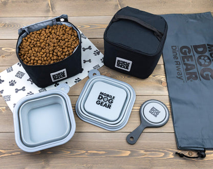 A collection of Mobile Dog Gear Dine Away® Food Sets, Large, Black by Mobile Dog Gear including two collapsible bowls, a food carrier filled with kibble, two closed storage bags, and a gray carrying bag, all displayed on a wooden surface.