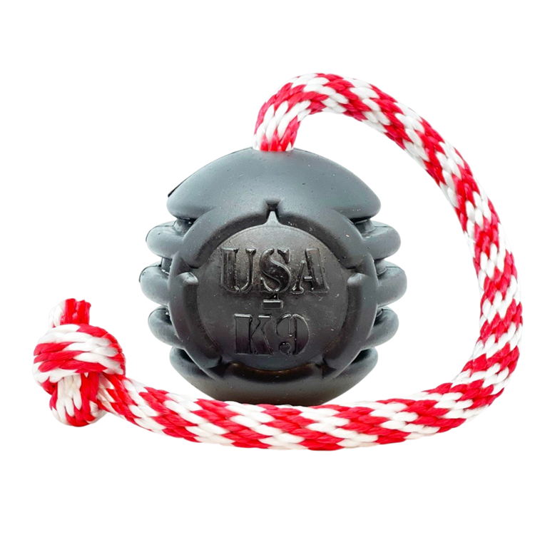 The SodaPup USA-K9 Magnum Black Stars and Stripes Ultra-Durable Rubber Chew, Tug, and Reward Toy features a grenade shape with a red and white braided rope. Made in the USA for tough chewers. Shown on a plain white background.