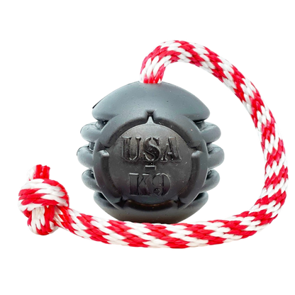 The SodaPup USA-K9 Magnum Black Stars and Stripes Ultra-Durable Rubber Chew, Tug, and Reward Toy features a grenade shape with a red and white braided rope. Made in the USA for tough chewers. Shown on a plain white background.