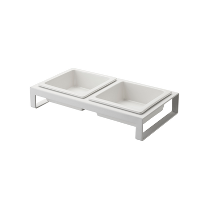 The Yamazaki Home Pet Food Bowl features a modern, white, elevated double feeder design with two square ceramic bowls resting on a sleek metal stand. This stylish setup is ideal for cats and small dogs, providing an elegant solution for their food and water requirements.