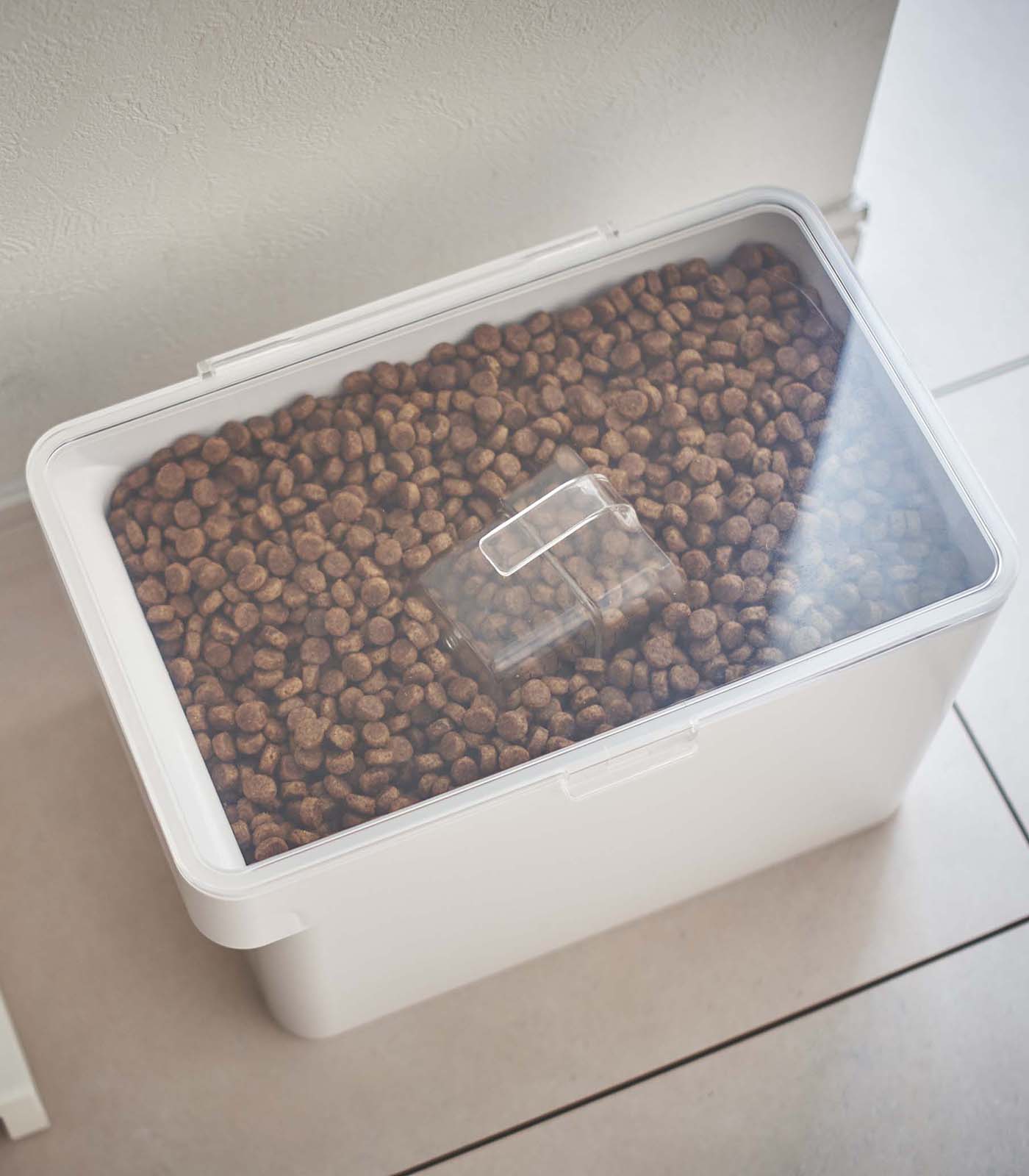 A white Yamazaki Home Airtight Pet Food Container - Three Sizes filled with kibble dog food on a tile floor.