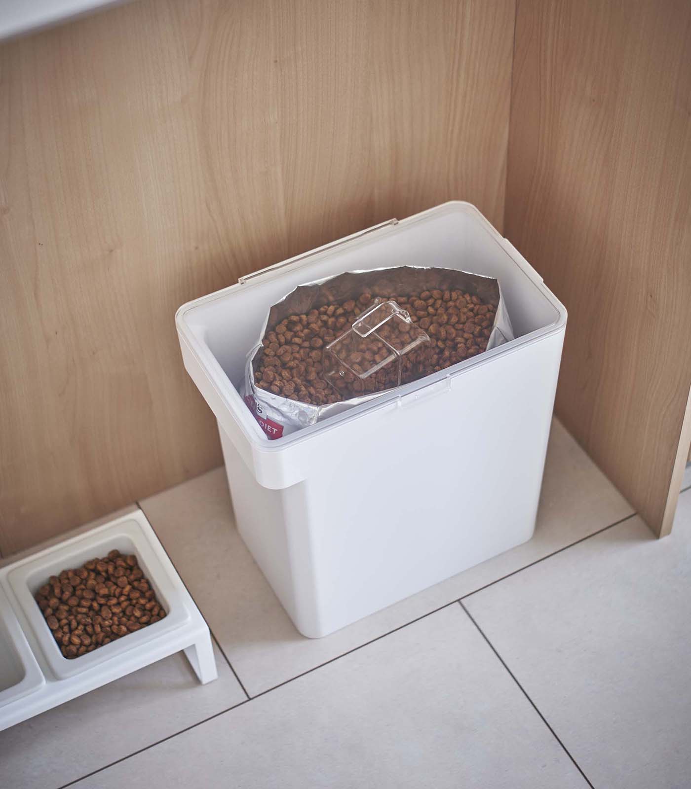 A white Yamazaki Home airtight pet food container filled with kibble on the floor next to a bowl of dog food.