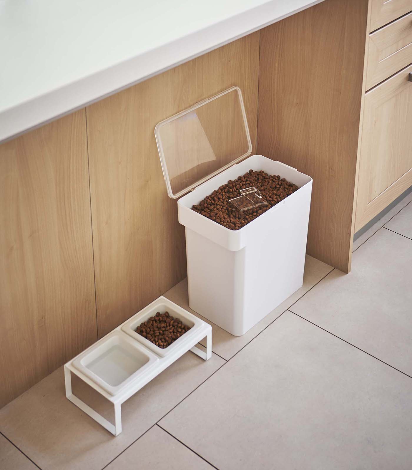 A large white Yamazaki Home airtight pet food container with a scoop on top stands beside two smaller trays filled with dry pet food, all set on a beige tile floor against a wooden kitchen cabinet.