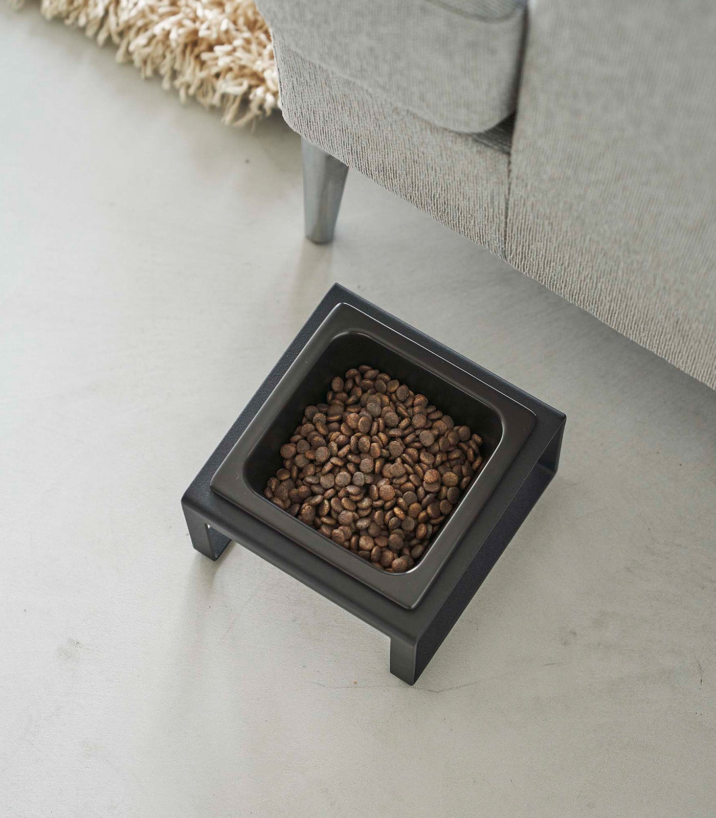 A Single Pet Food Bowl - Two Styles - Steel + Ceramic by Yamazaki Home on the floor next to a couch.