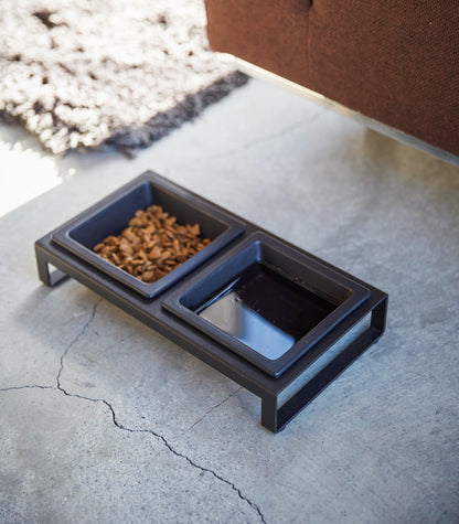 A small black Pet Food Bowl made of ceramic on the floor next to a couch. Brand Name: Yamazaki Home.