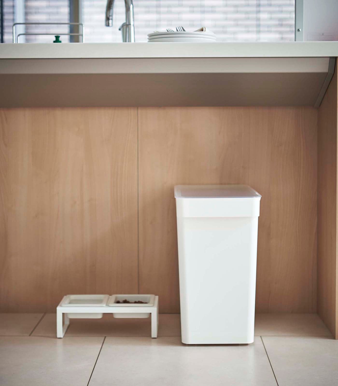 A Yamazaki Home Rolling Airtight Pet Food Container (25 Lbs.), serving as an airtight kibble storage solution, sits on a wooden floor in a kitchen.