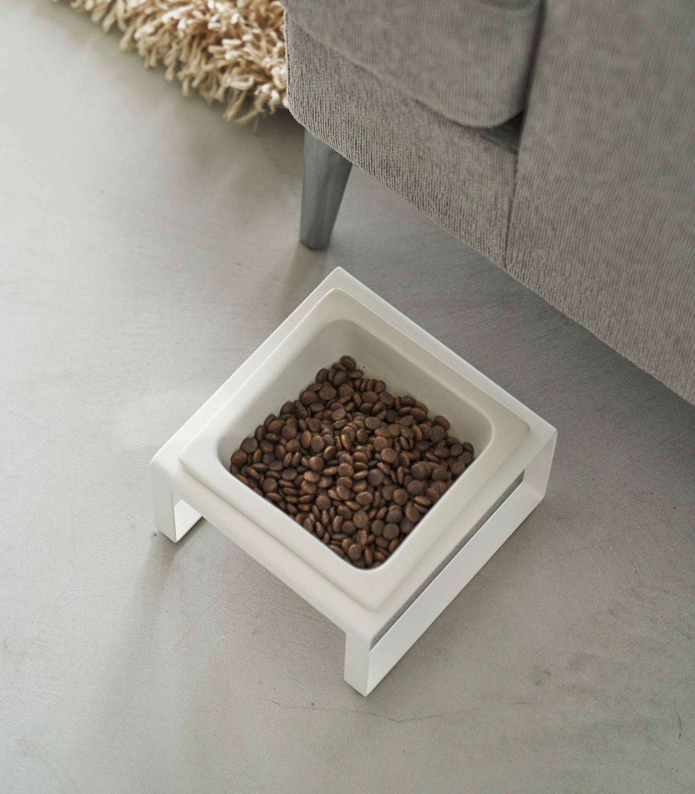 A Single Pet Food Bowl - Two Styles - Steel + Ceramic by Yamazaki Home on the floor next to a couch.