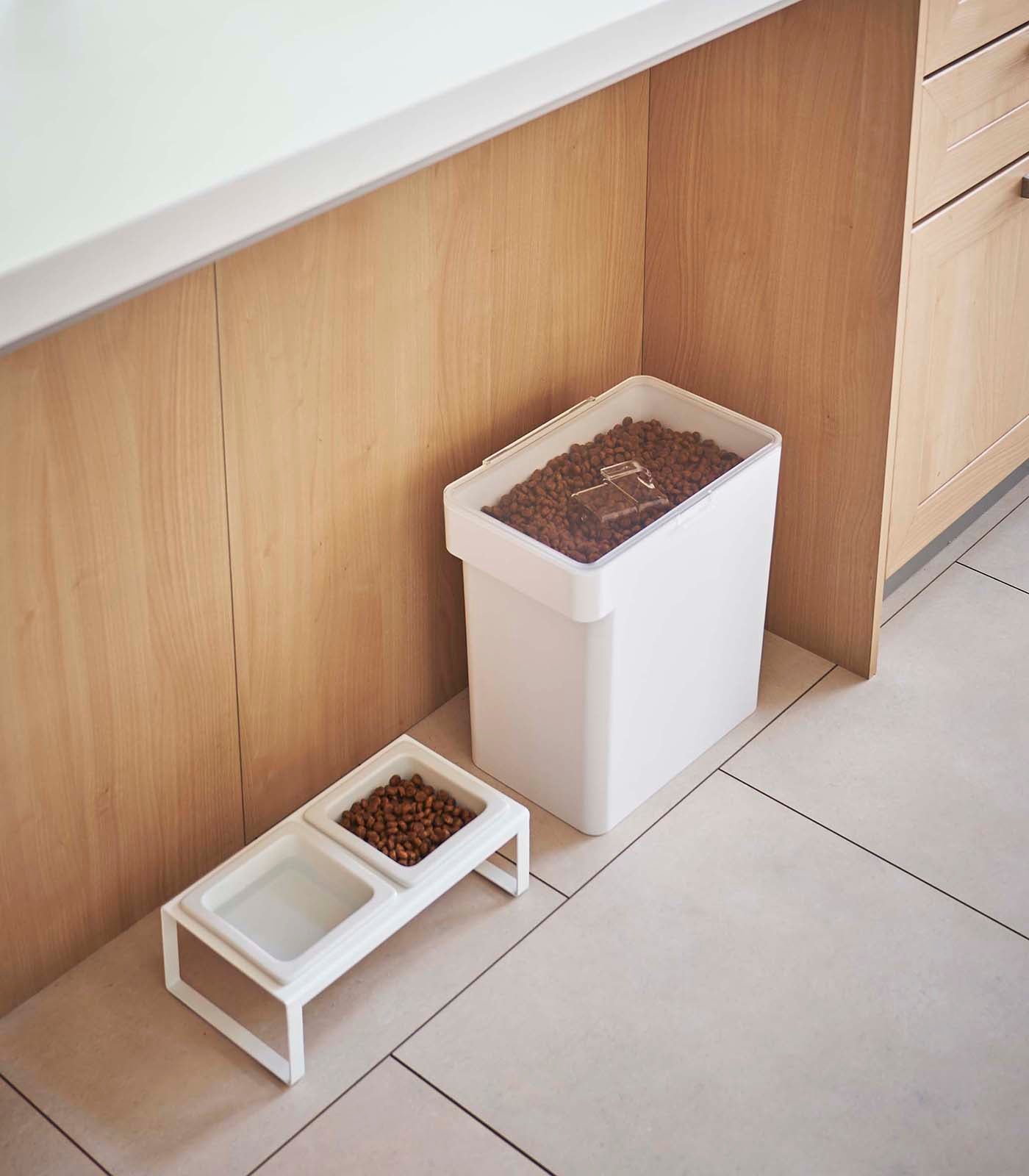 A Yamazaki Home Airtight Pet Food Container - Three Sizes next to a two-section feeding station with kibble, positioned against a kitchen cabinet on a tiled floor.