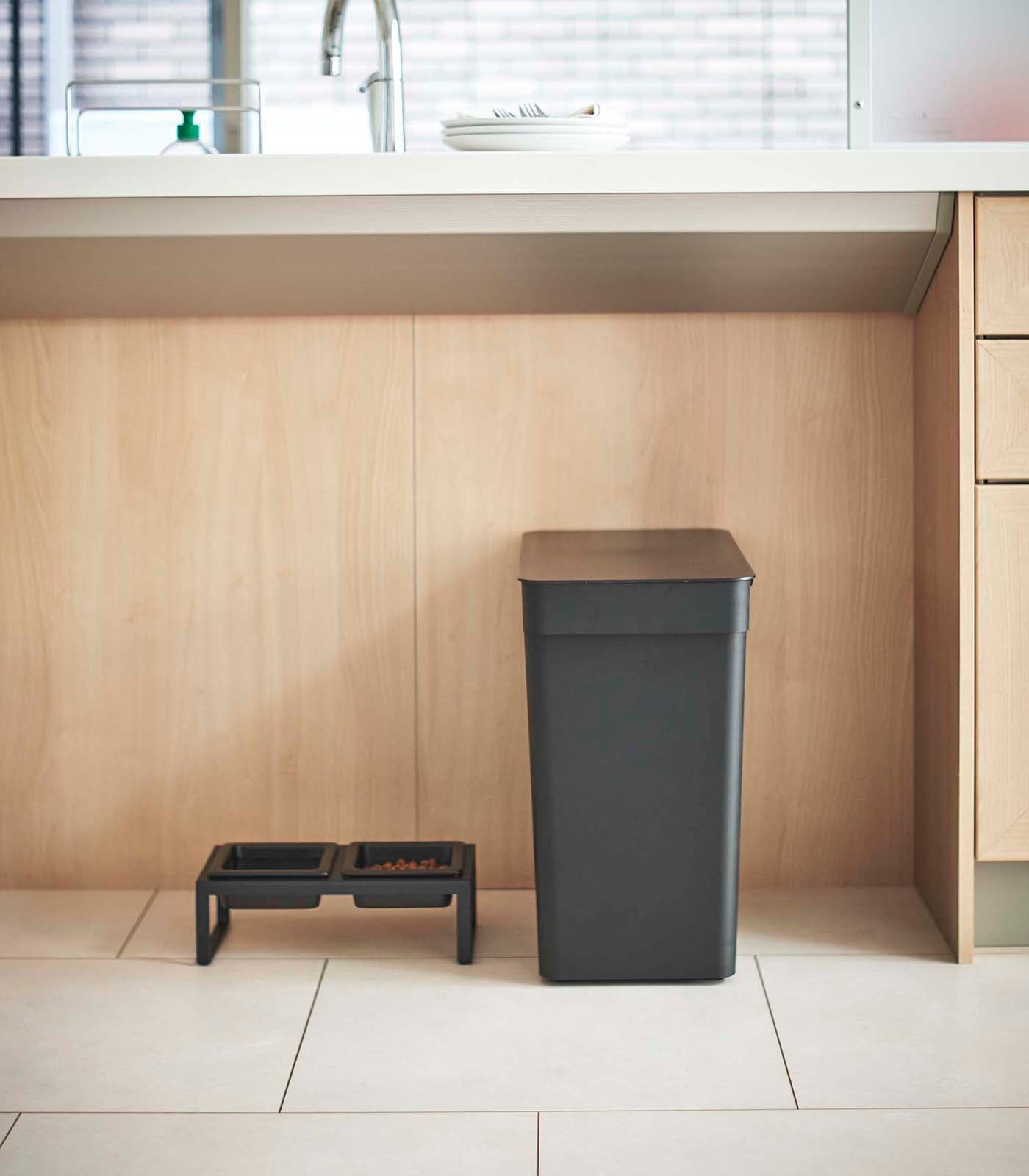 A Yamazaki Home Rolling Airtight Pet Food Container (25 Lbs.) sits on a counter in a kitchen. On the other side of the counter, an airtight kibble storage solution from Yamazaki Home keeps dry pet food fresh.