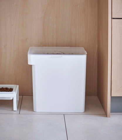 A Yamazaki Home white trash bin placed on a light wood floor against a matching light wood cabinet, with a small tray containing Yamazaki Home Airtight Pet Food Container - Three Sizes on the left side.