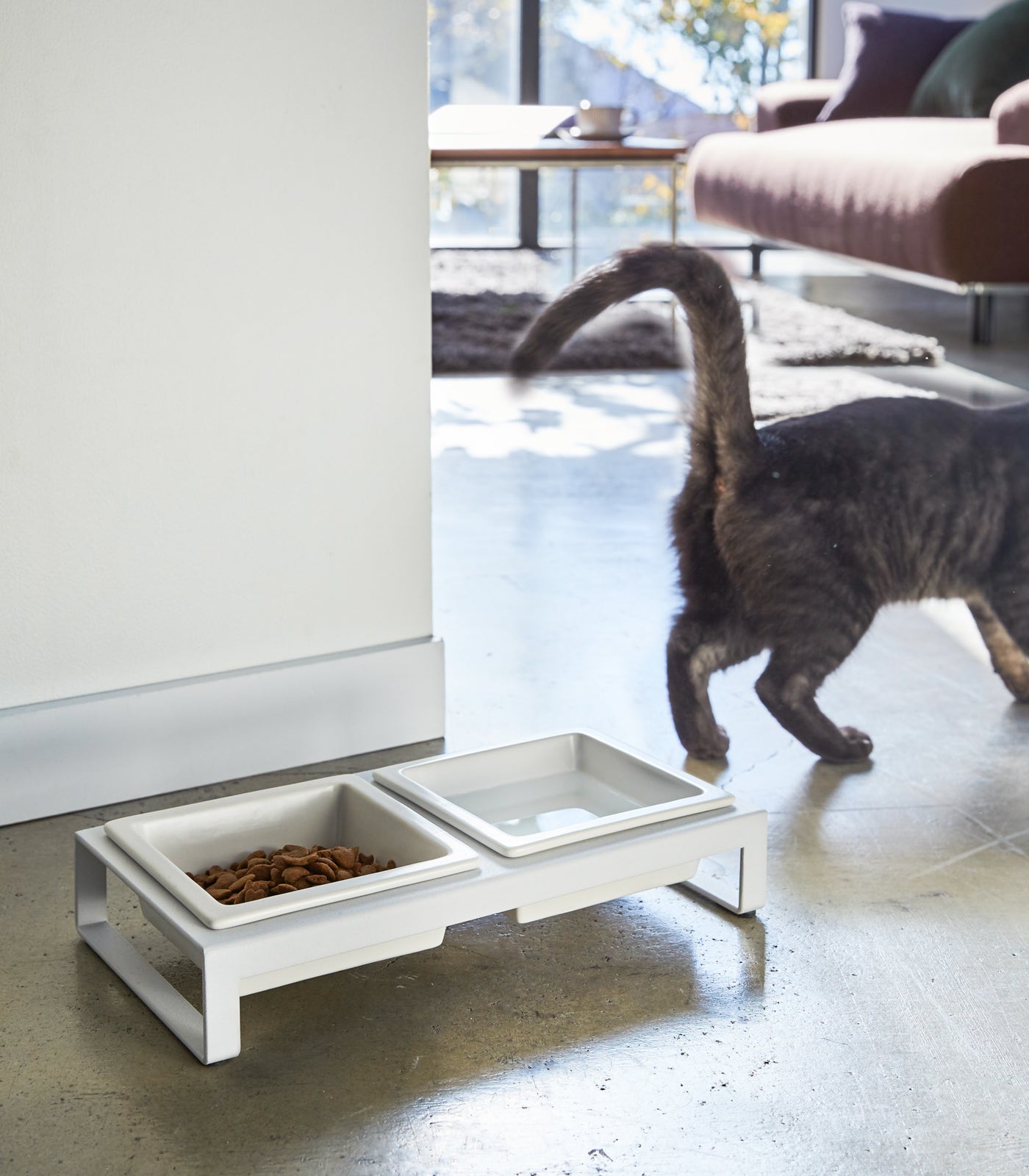 A cat walking away from a two-section Yamazaki Home ceramic pet food bowl placed on a gray floor.