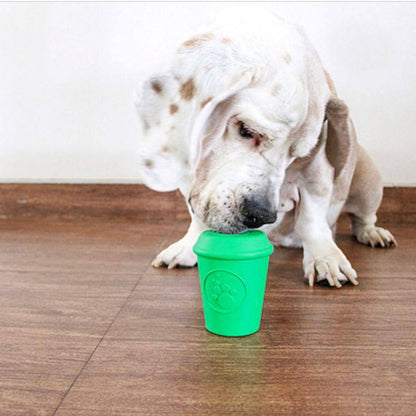 A white and brown dog with long ears curiously observes a small, coffee cup-shaped toy from the SodaPup brand, officially known as the Coffee Cup EDispener Durable Rubber Chew Toy and Treat Dispenser, featuring a paw print design, resting on the wooden floor against a plain white wall backdrop.