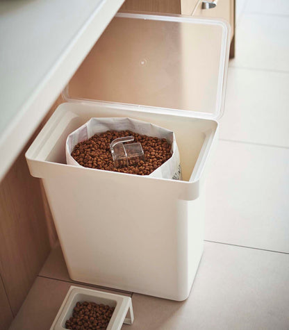 A white Yamazaki Home Rolling Airtight Pet Food Container (25 Lbs.) with a lid on it next to a rolling measured cup.