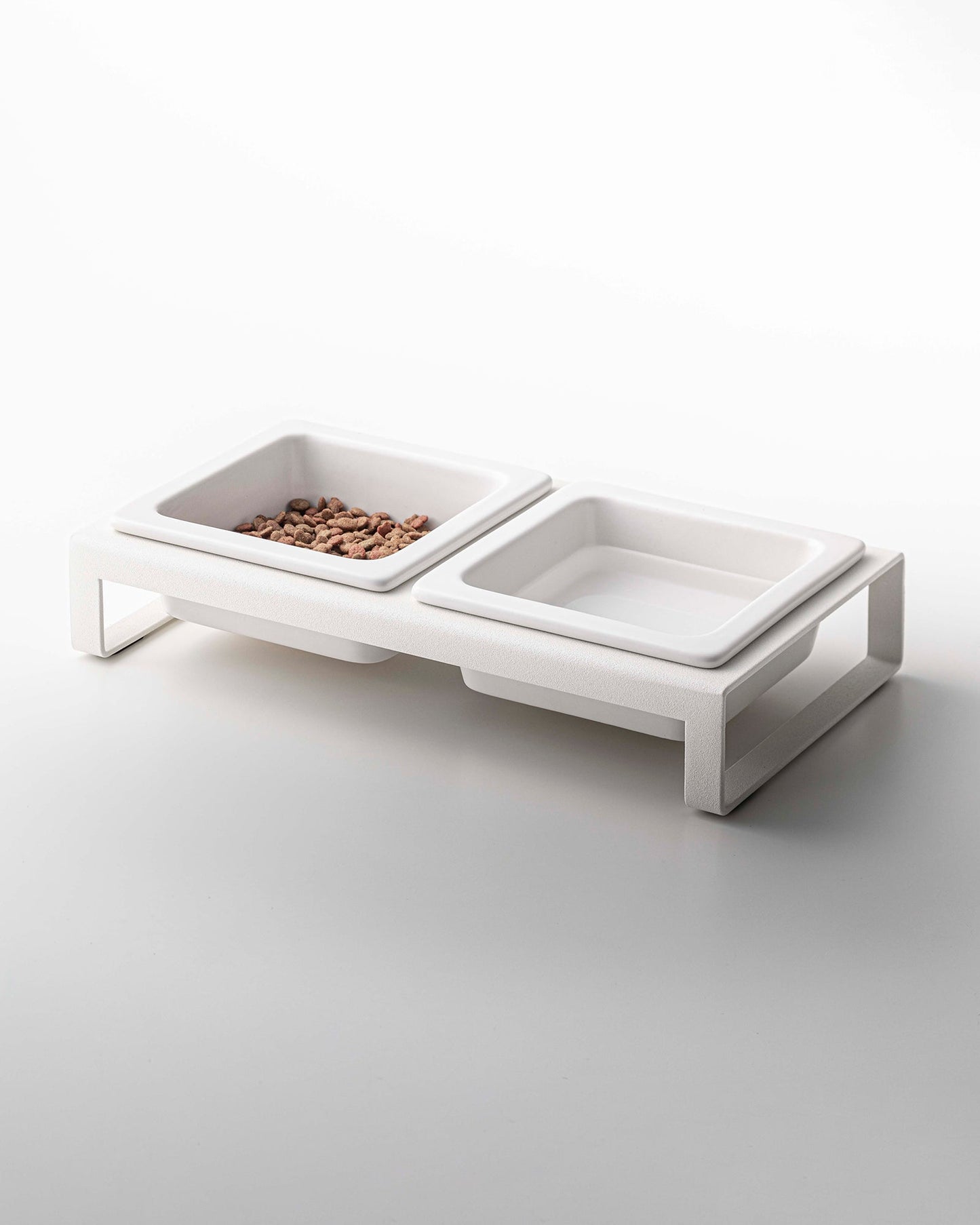 Elevated ceramic Yamazaki Home pet food bowls with one bowl containing food and the other empty, designed for small dogs, against a white background.