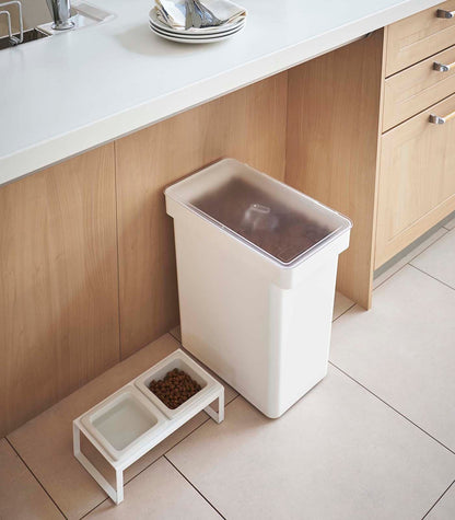 A kitchen with a white trash can and a bowl of food, showcasing the Yamazaki Home Rolling Airtight Pet Food Container (25 Lbs.), an airtight kibble storage solution.