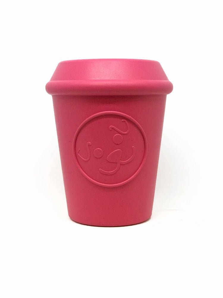 A pink reusable cup from SodaPup known as the Coffee Cup EDispener Durable Rubber Chew Toy and Treat Dispenser stands upright, resembling a coffee cup toy. Its front is adorned with an embossed circular logo that enhances its sleek design. The plain white background accentuates the cup's vibrant color.