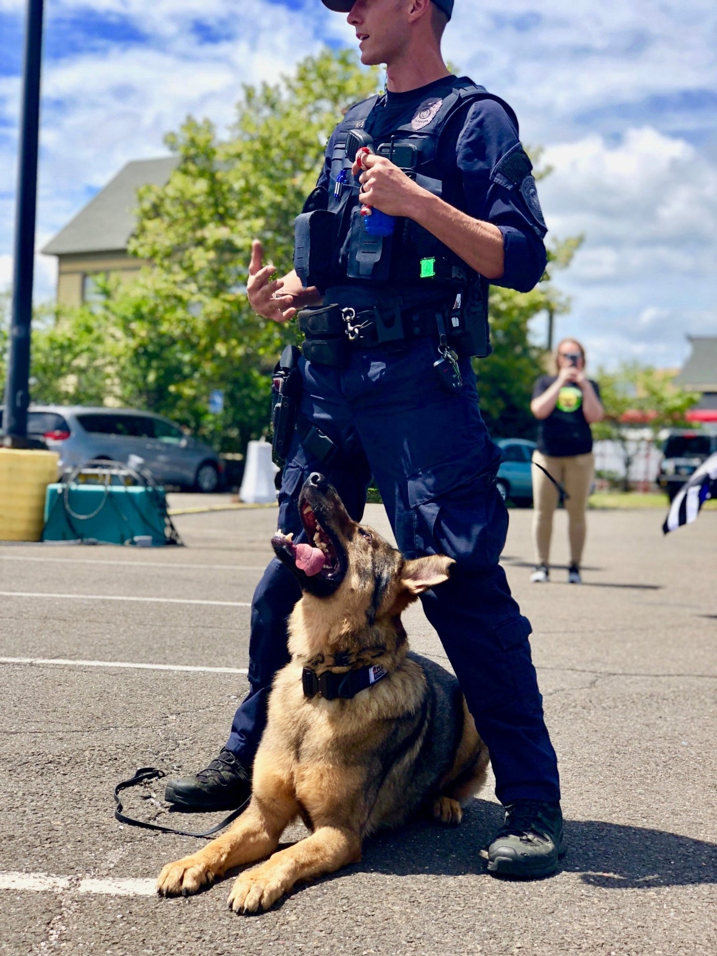 A police officer with a german shepherd equipped with a USA-K9 Magnum Grenade Durable Rubber Chew Toy, Treat Dispenser, Reward Toy, Tug Toy, and Retrieving Toy - Black Magnum from SodaPup patrols a parking lot.