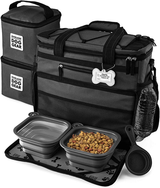 A black and gray Mobile Dog Gear Rolling Week Away Dog Travel Bag set including a main compartment, side pouch, two collapsible feeding bowls with dog food, and a feeding mat. The items are branded with "Overland Dog Gear.