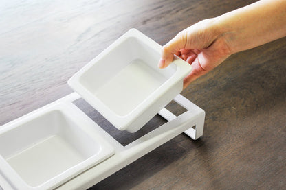 A hand arranging white ceramic bowls, including a Yamazaki Home pet food bowl for small dogs, on a wooden table.