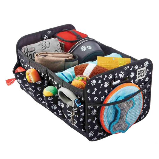 A black and white paw-print Mobile Dog Gear Dogssentials Collapsible Storage Organizer filled with various pet items, including a folded blanket, dog bowls, toys, a Frisbee, grooming tools, and napkins. This collapsible organizer features multiple compartments and pockets for keeping items neatly arranged and accessible.