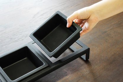 A person holding a small black tray above a set of ceramic Yamazaki Home pet food bowls on a wooden surface.